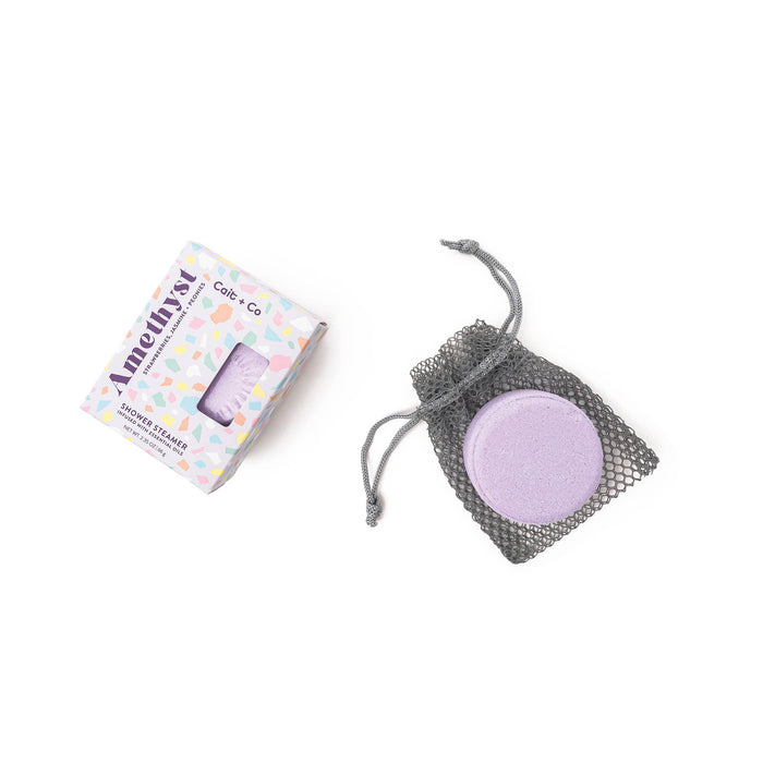 A shower steamer bag with a purple steamer visible through the mesh, alongside its packaging box with 'Cait + Co. Gem Shower Steamer - Amethyst' written on it.